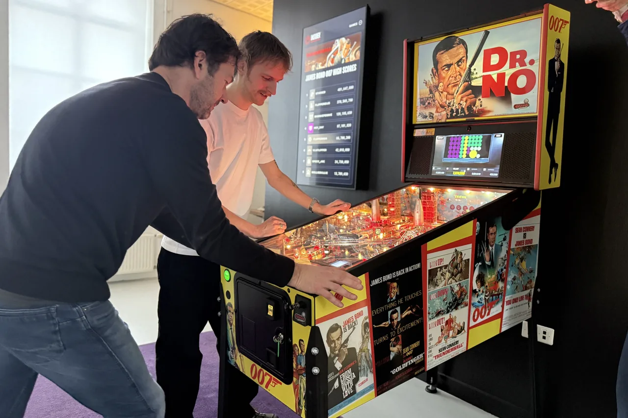 Twee collega's van Heyalo spelen op de James Bond 007 flipperkast, met het realtime leaderboard op het scherm ernaast