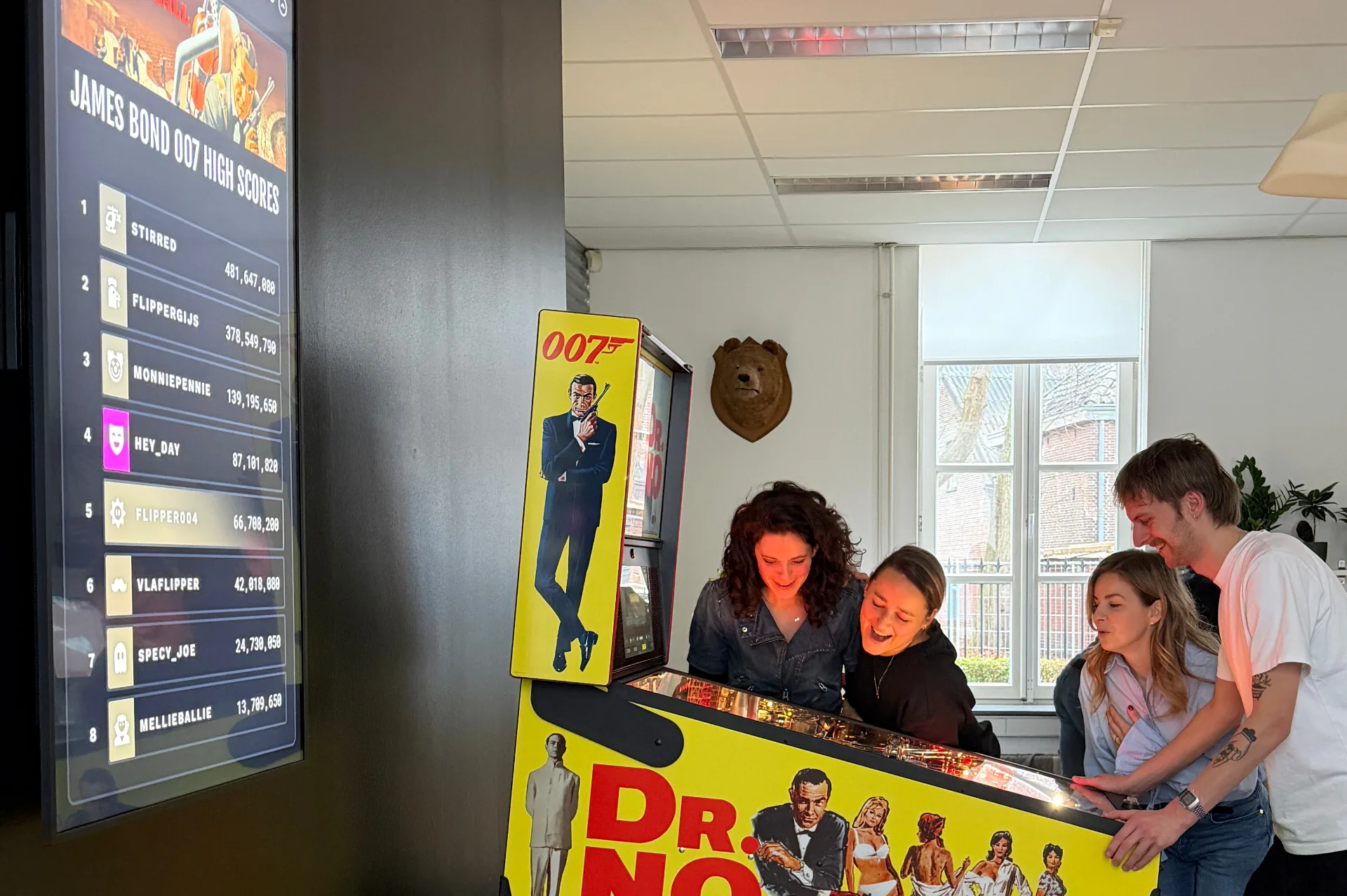 Vier collega's lachend rond de Dr. No-flipperkast, met de James Bond 007 high scores leaderboard op het scherm ernaast